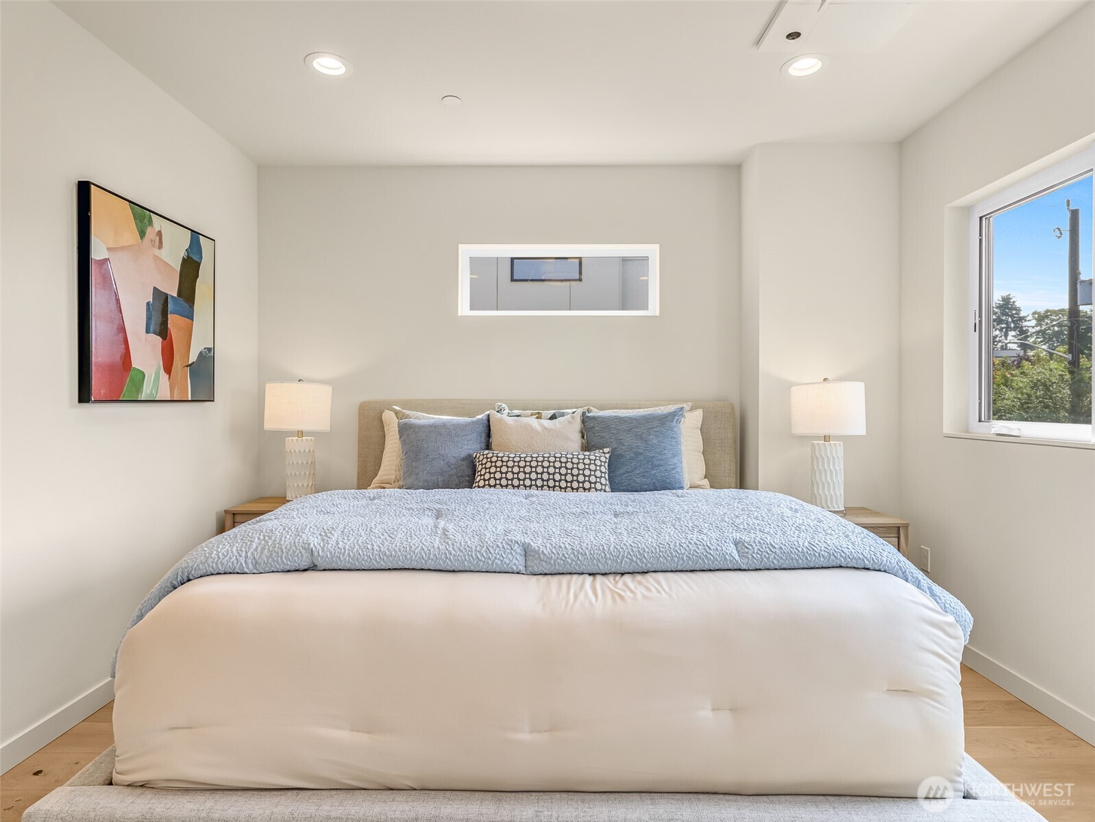 Staged bedroom with a large white bed, blue pillows, matching lamps, and a window overlooking trees.