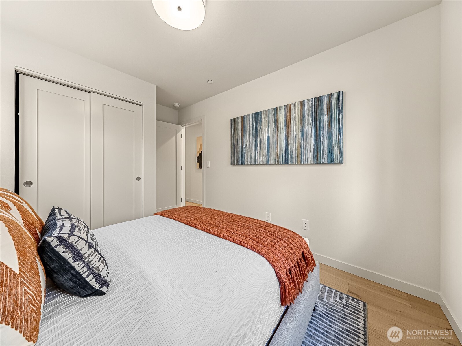 Staged bedroom with a white bed, rust-colored throw, blue abstract artwork, and sliding-door closet.
