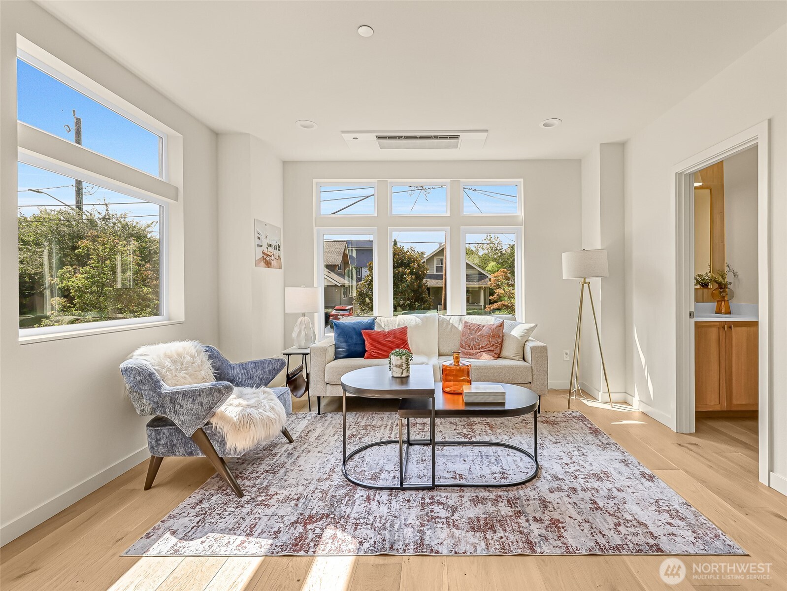 Staged living room with large windows, a beige sofa, blue chair, and light wood floors.
