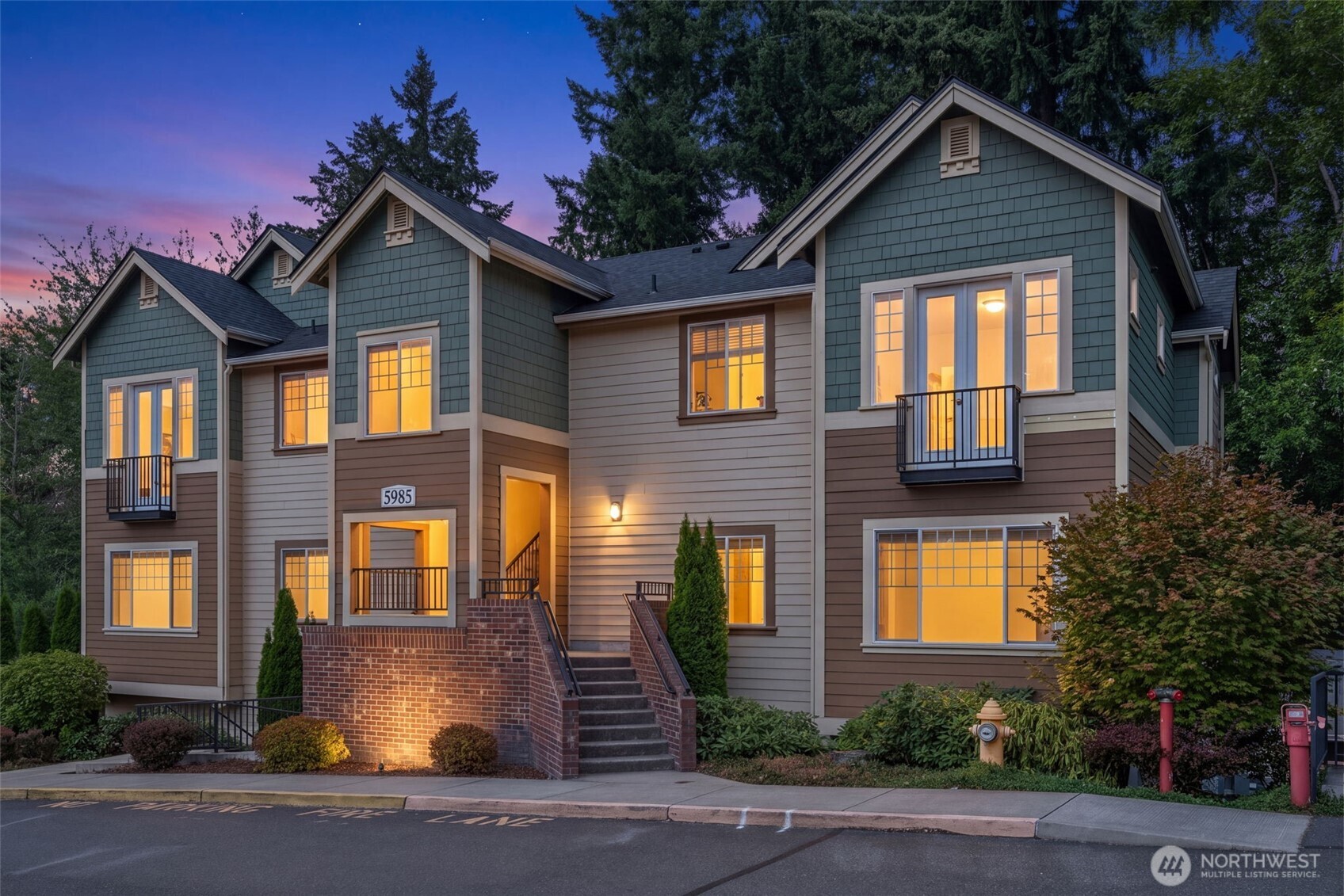 Multi-unit residential building with brick entry walls, balconies, and glowing windows at dusk.