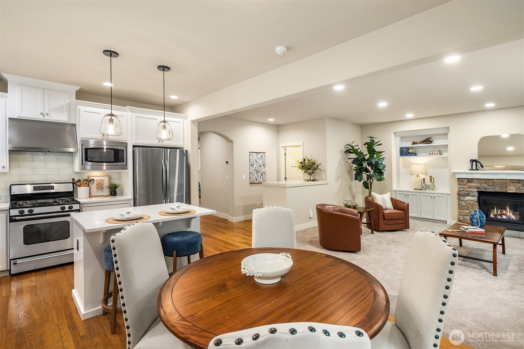 Open-concept kitchen and living room with white cabinets, a round wood dining table, and stone fireplace.