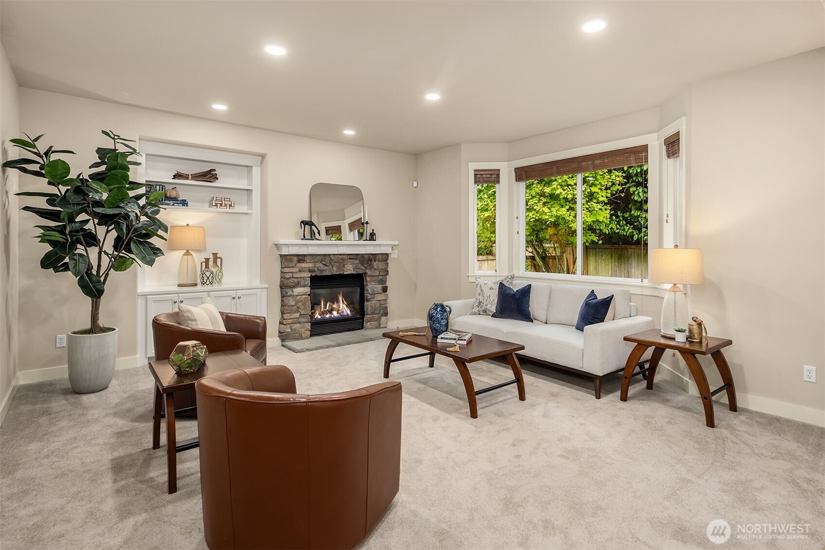 Staged living room with a stone fireplace, built-in shelving, and a cream sofa beside a leafy window.
