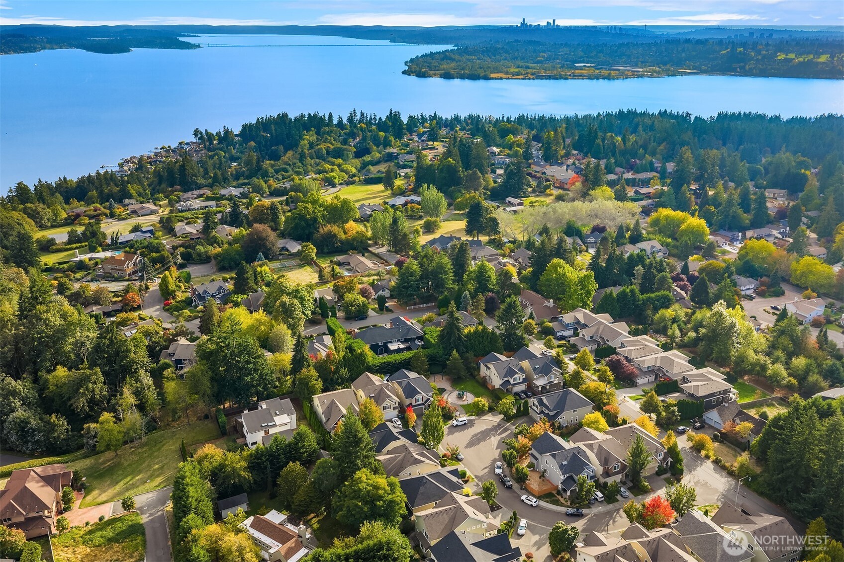 Aerial view of Kirkland homes and autumn trees beside a broad lake, with a distant city skyline.