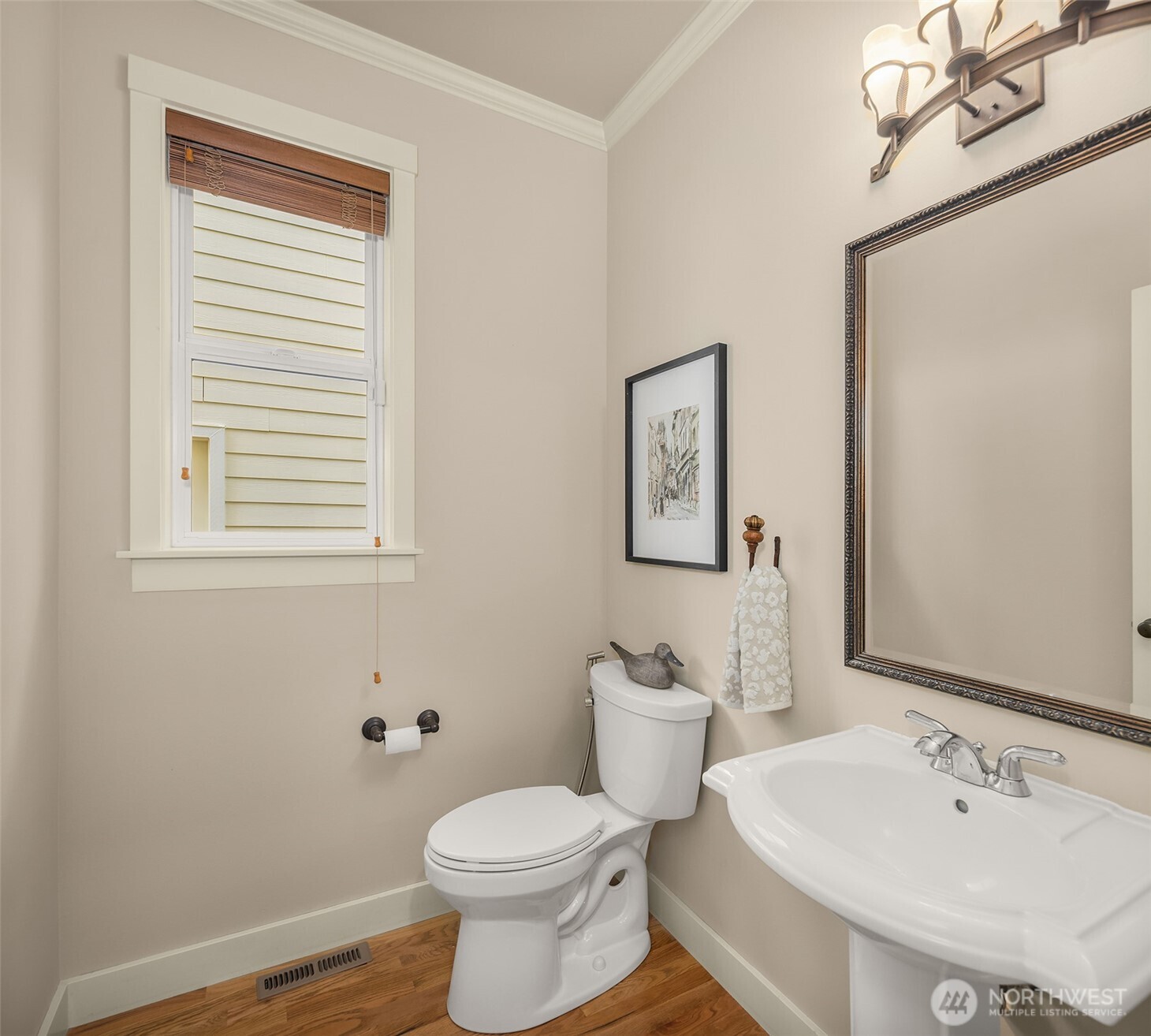 Bathroom with a white pedestal sink, toilet, framed mirror, and wood flooring.