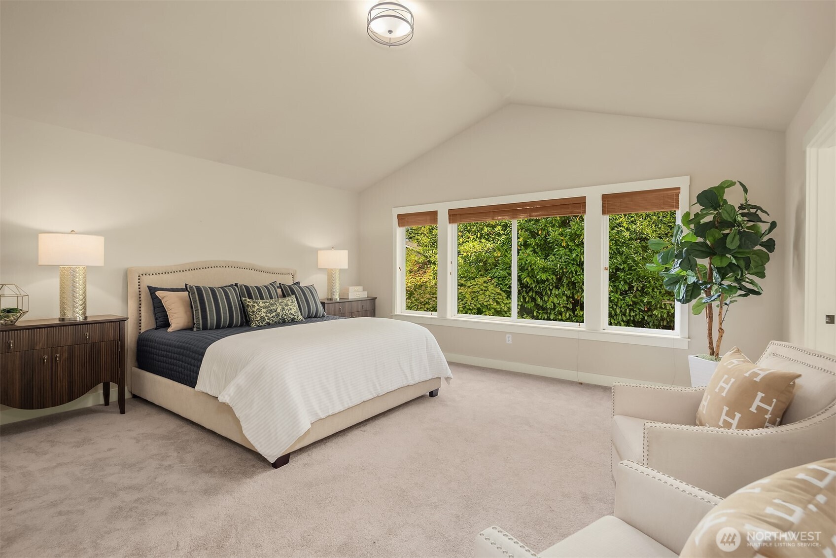 Staged bedroom with a large bed, vaulted ceiling, and wide windows overlooking greenery.