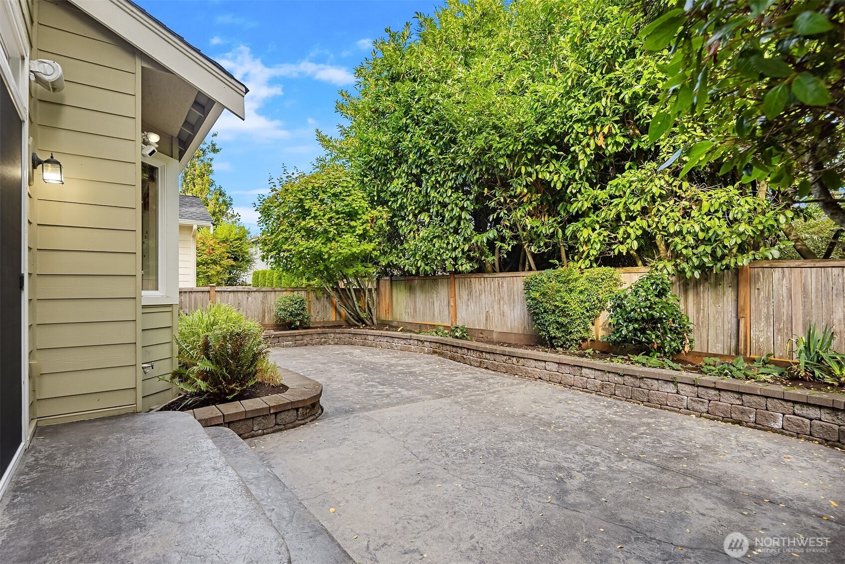 Paved patio beside the house, surrounded by raised garden beds, mature greenery, and a wood privacy fence.