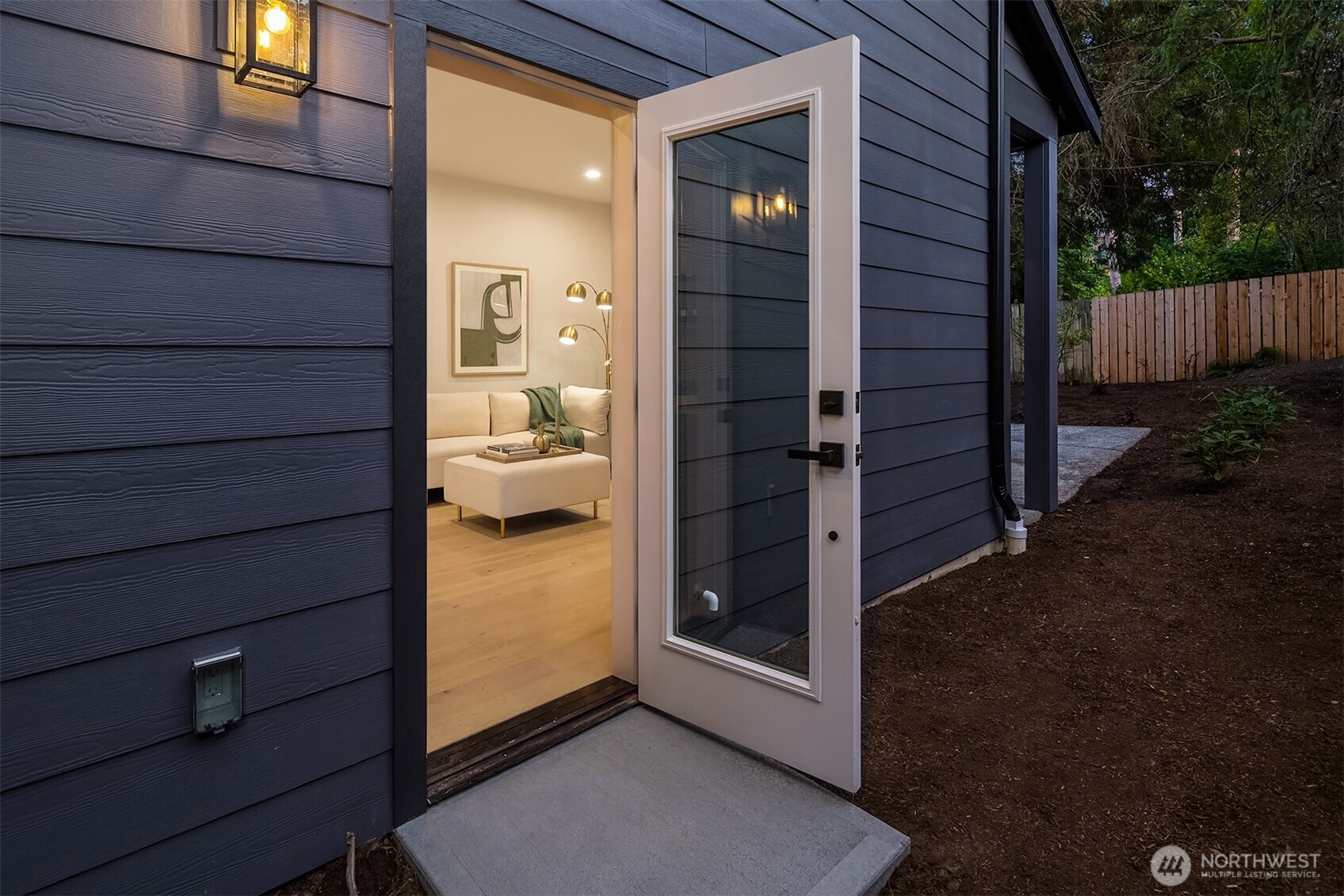 Open front door in dark blue siding reveals a warmly lit living room and staged seating.