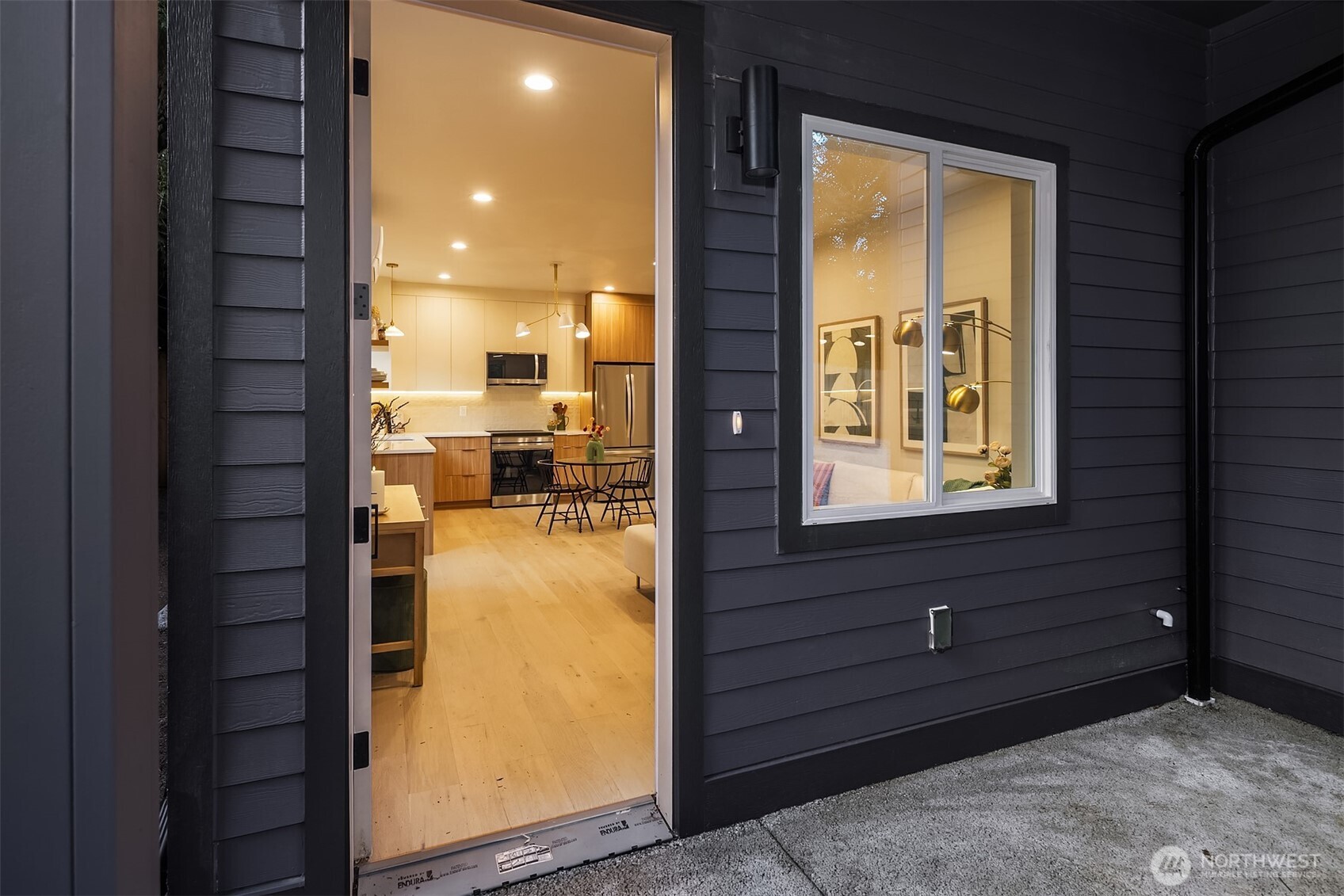 Open doorway in dark siding reveals a warmly lit kitchen with wood cabinetry and a dining table.