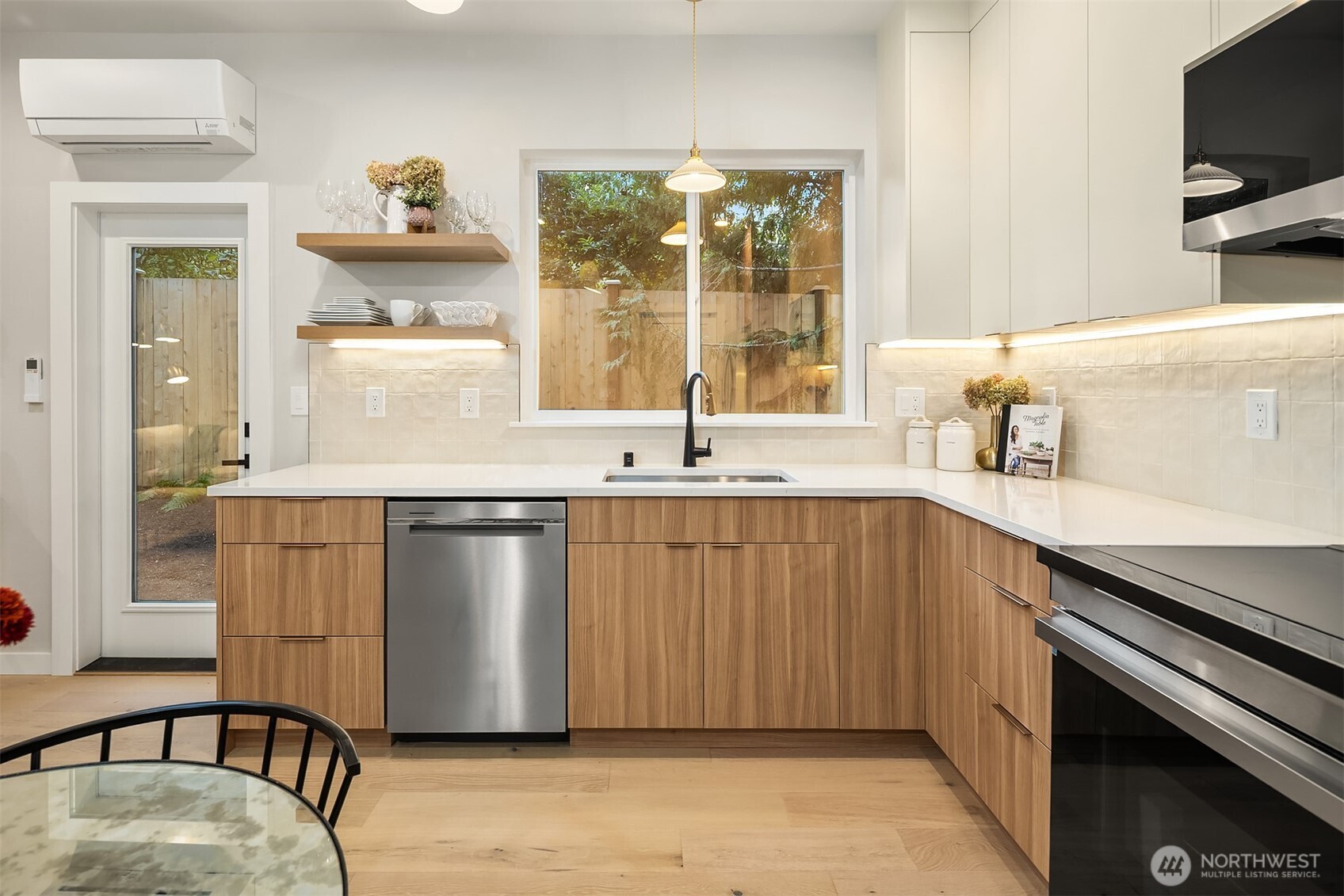 Modern kitchen with light wood cabinets, white counters, stainless appliances, and a large window overlooking a fenced yard.