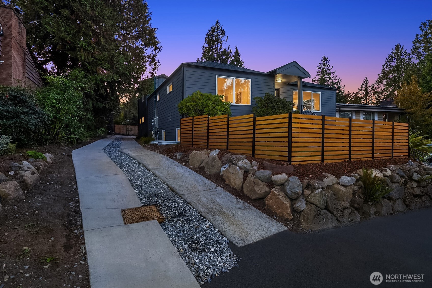 Modern dark-sided home at dusk with a wood privacy fence and curved concrete walkway.
