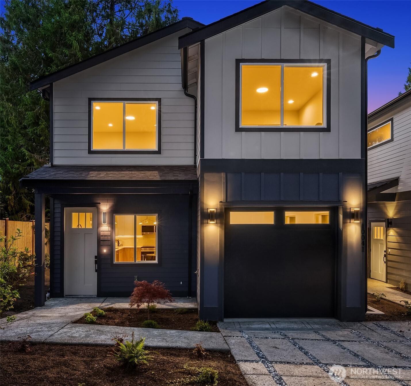 Modern two-story home with dark siding, glowing windows, and a wide garage at dusk.