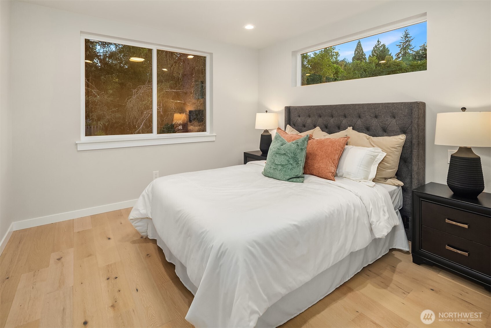 Staged bedroom with a white bed, charcoal tufted headboard, and windows overlooking trees.
