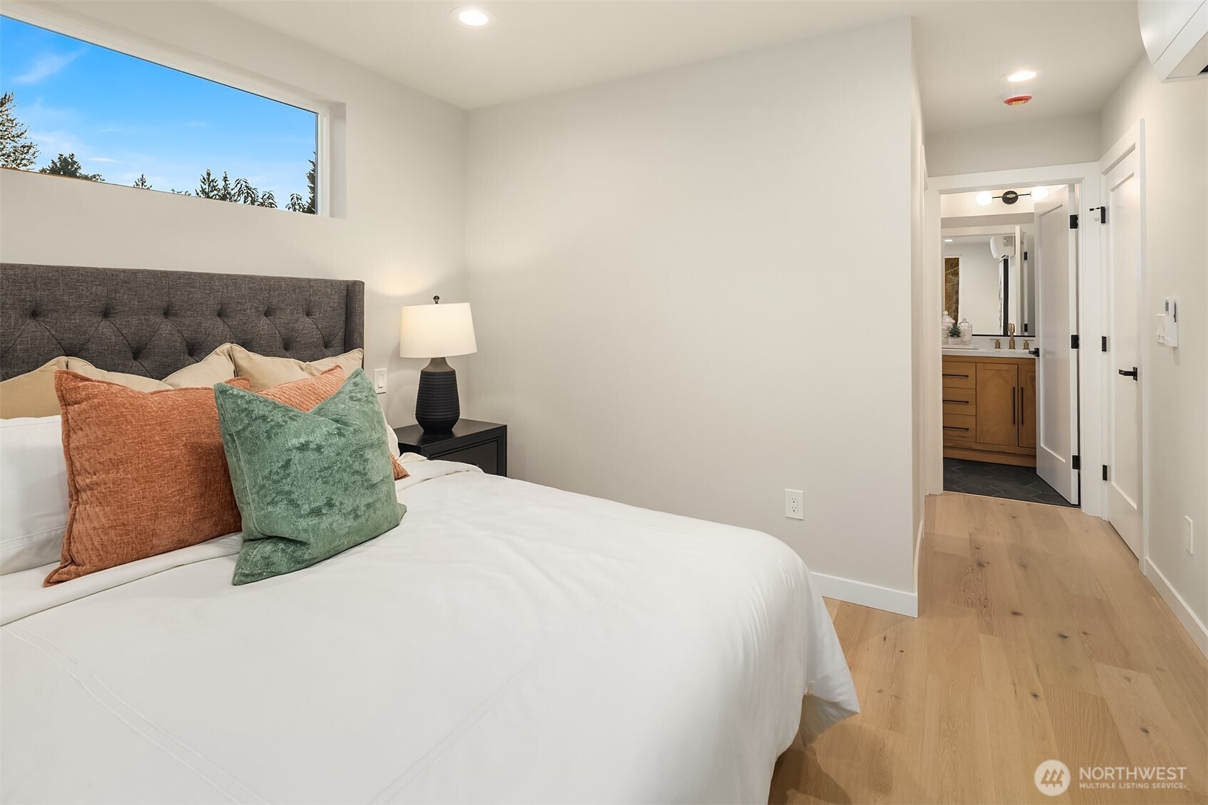 Staged bedroom with a white bed, colorful accent pillows, and an open doorway to a bathroom.