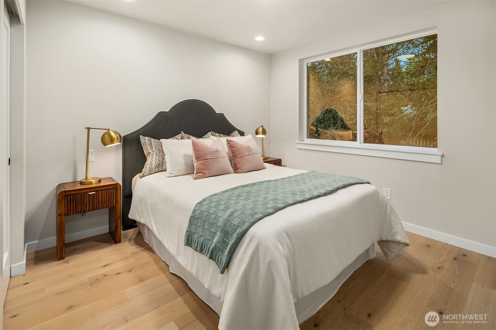 Staged bedroom with a dark curved headboard, sage-green bed throw, and large window overlooking autumn trees.
