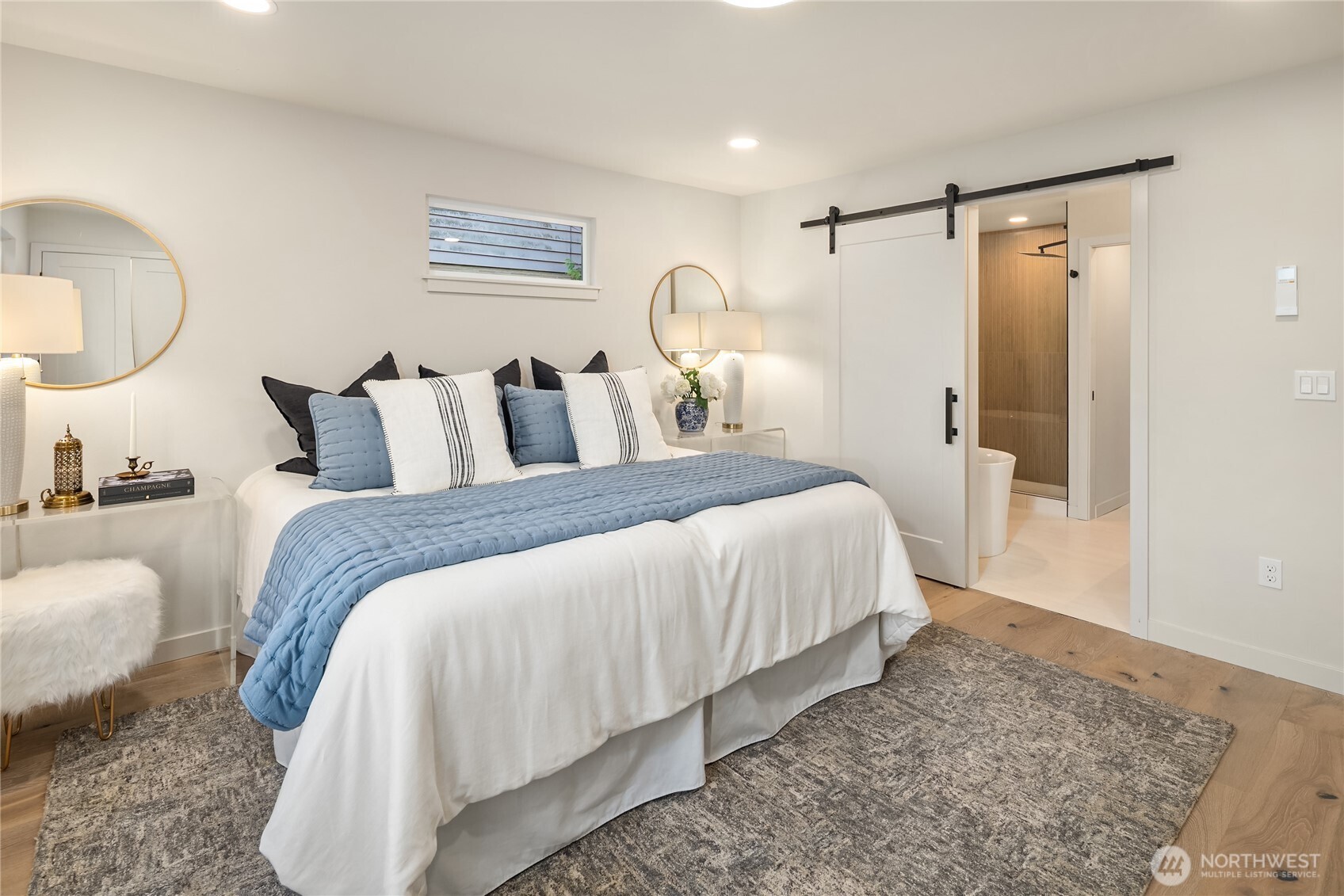 Staged bedroom with a white bed, pale blue quilt, and sliding barn door to the bathroom.