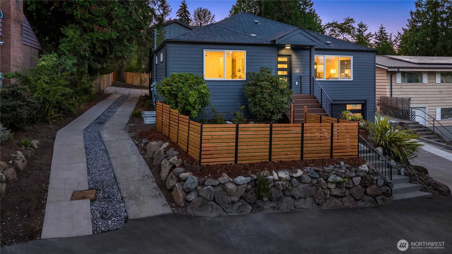 Blue-sided home with warmly lit windows, a wood fence, and a stone retaining wall at dusk.