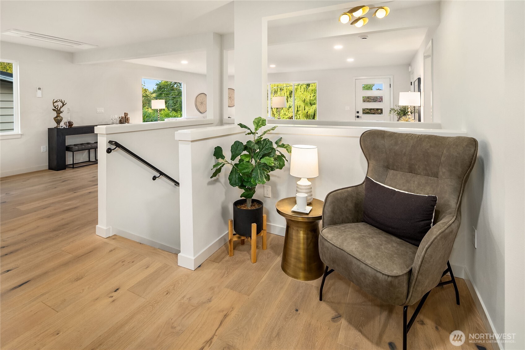 Furnished sitting area with a gray armchair, brass side table, and potted plant beside an open entry.