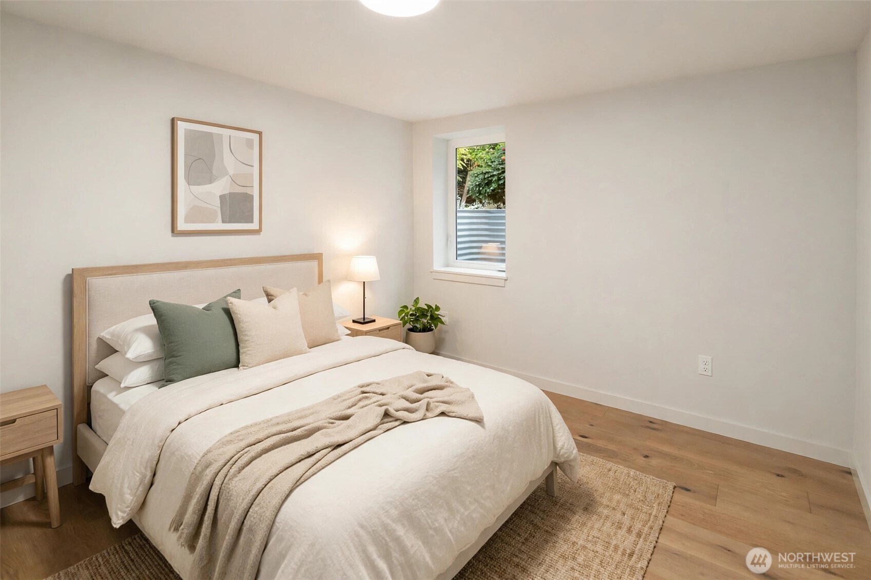 Staged bedroom with white bedding, green accent pillows, light wood furniture, and a window overlooking greenery.
