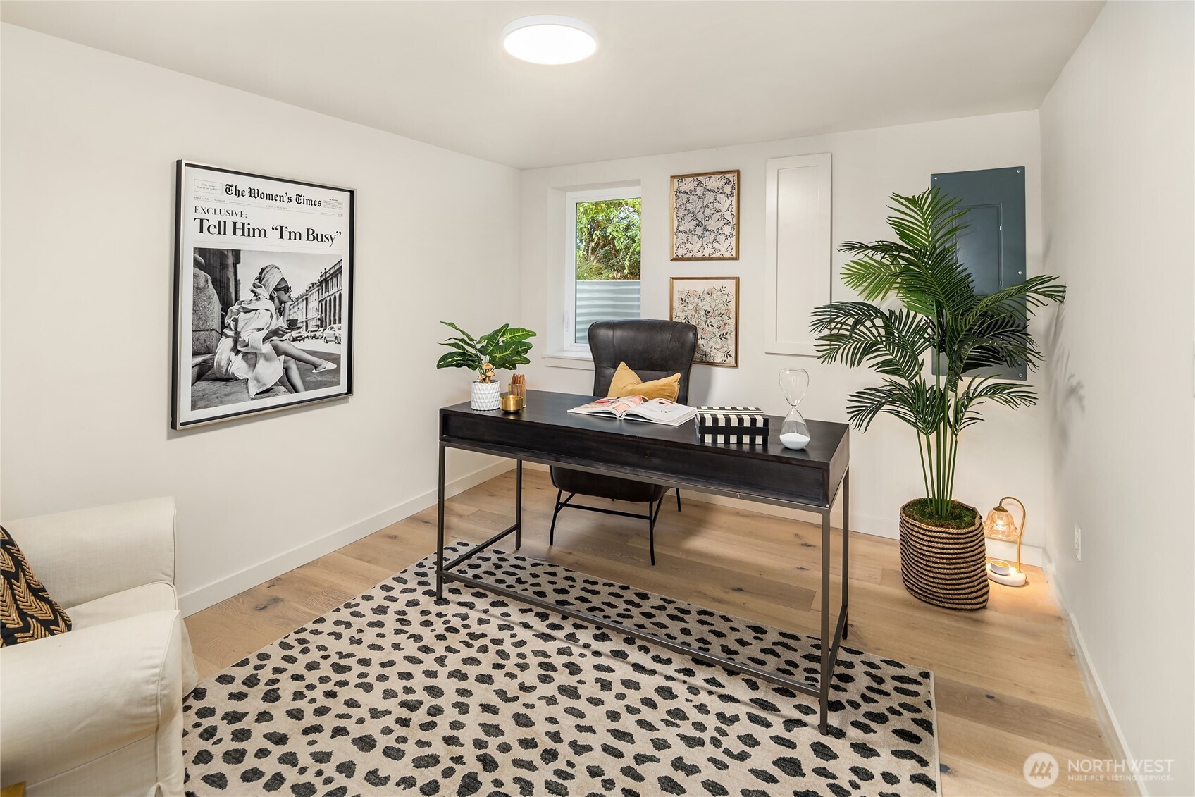 Staged home office with a dark desk, leather chair, patterned rug, and large potted palm.