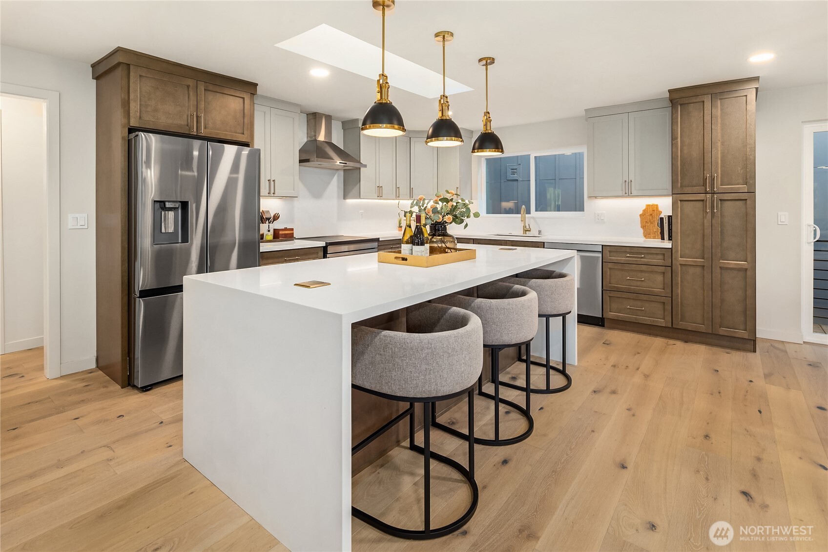 Modern kitchen with a large white island, upholstered stools, dark wood cabinetry, and stainless steel appliances.