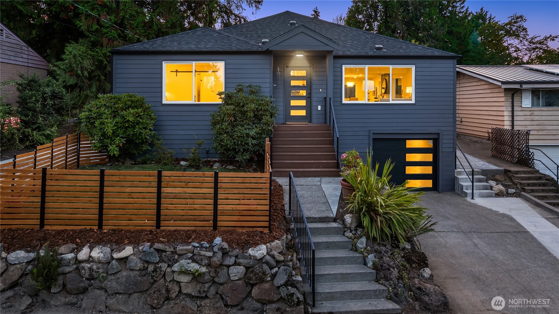 Blue-gray single-story home with warmly lit windows, front steps, wood fencing, and a stone retaining wall.