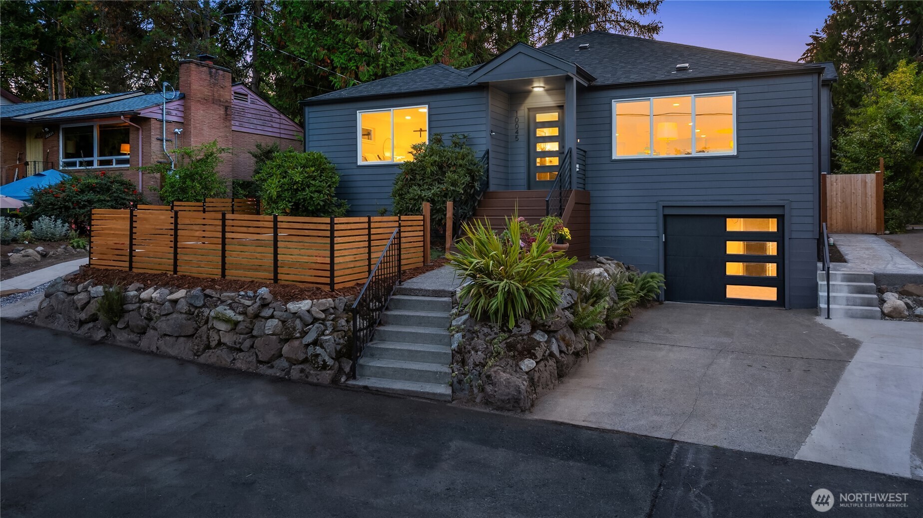 Dark blue-gray house with warmly lit windows, a single-car garage, stone landscaping, and a wooden front fence.
