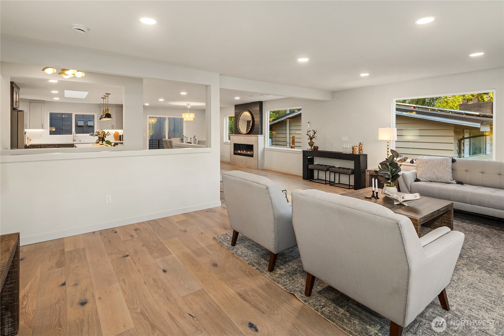 Open-plan living room with pale seating, wood floors, fireplace, and kitchen beyond.