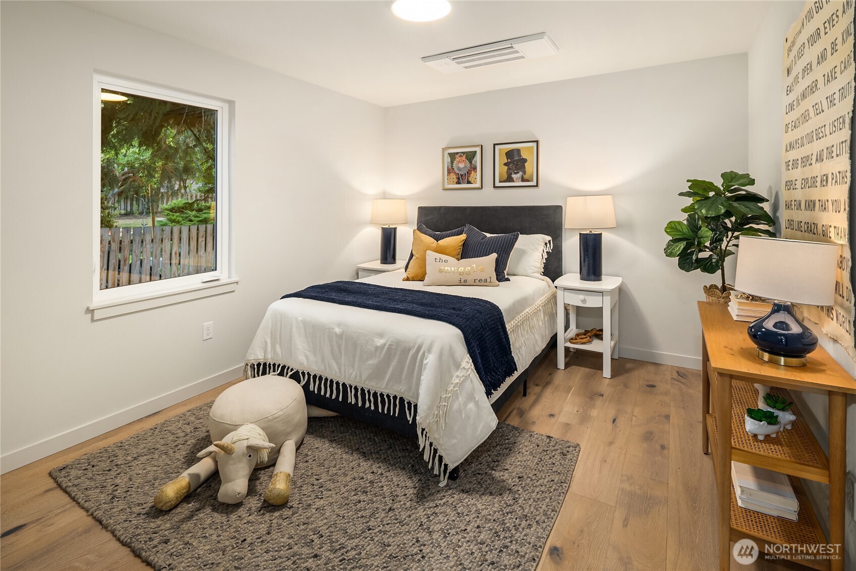 Staged bedroom with white and navy bedding, a leafy yard visible through the window, and a woven rug.
