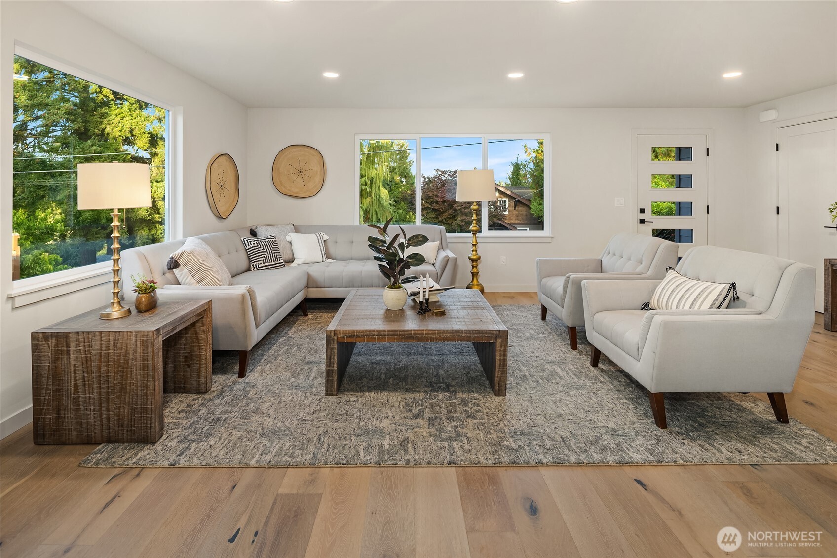 Staged living room with a gray sectional, matching armchairs, and wide windows overlooking trees.