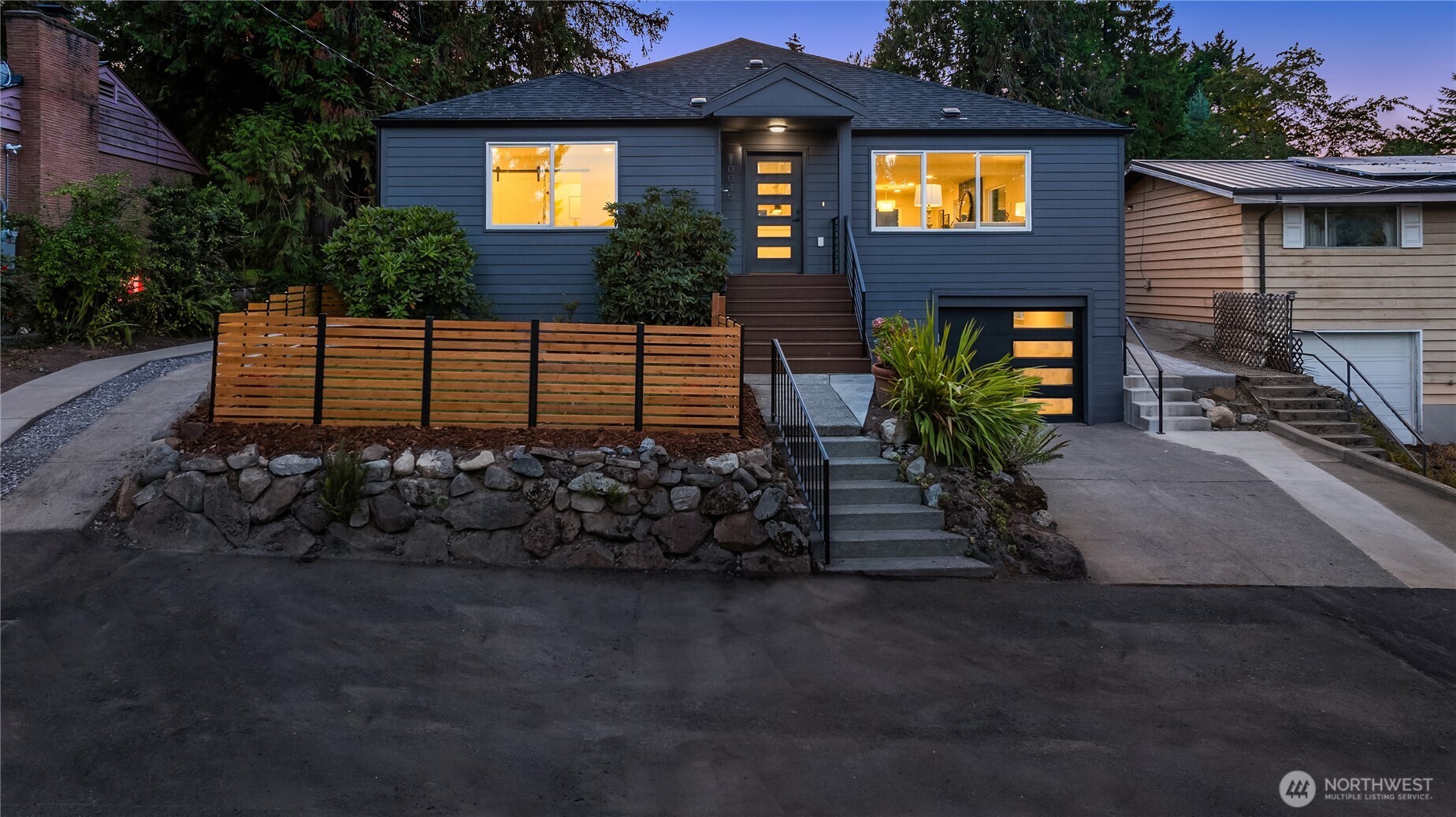 Dark blue Seattle home at dusk with warmly lit windows, front steps, and a wood-slat fence.
