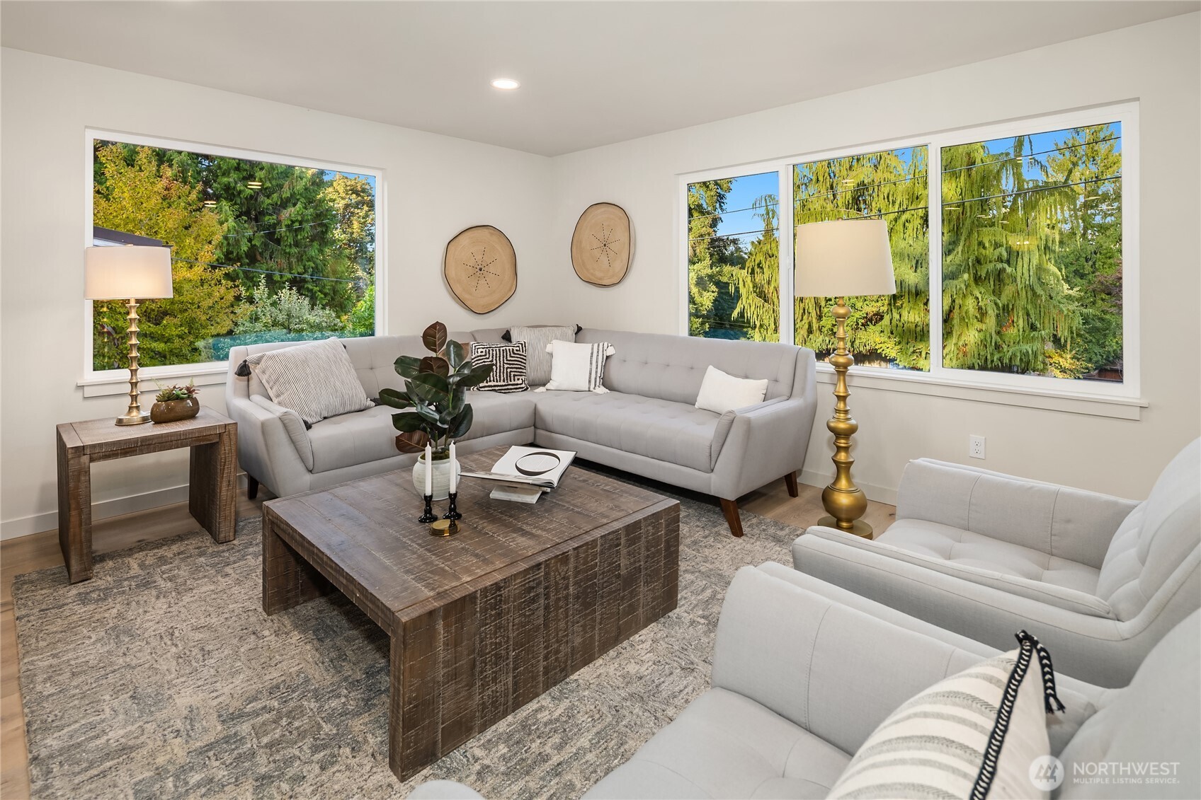 Living room with a gray sectional, rustic coffee table, and large windows overlooking leafy trees.