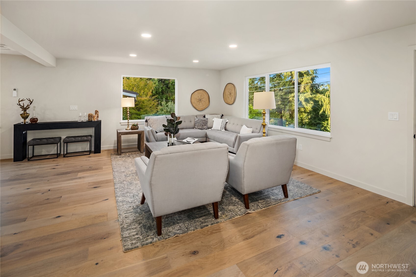 Staged living room with a gray sectional, wood floors, and large windows overlooking trees.