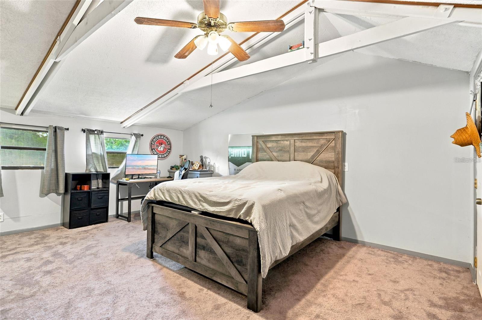 Bedroom with a rustic wood bed, exposed ceiling beams, and a desk beside the windows.