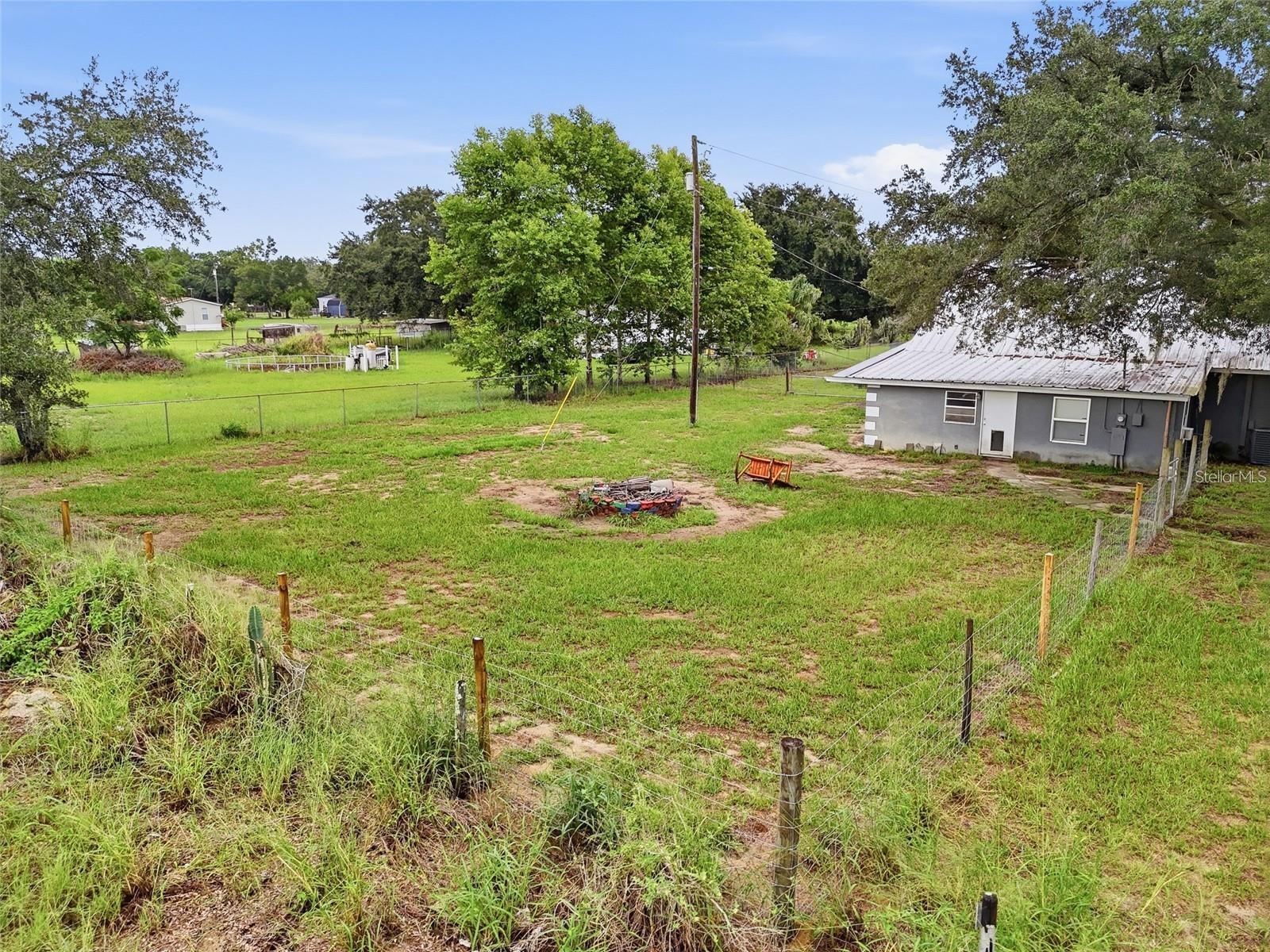 Fenced grassy lot with mature trees and a gray outbuilding with a metal roof.