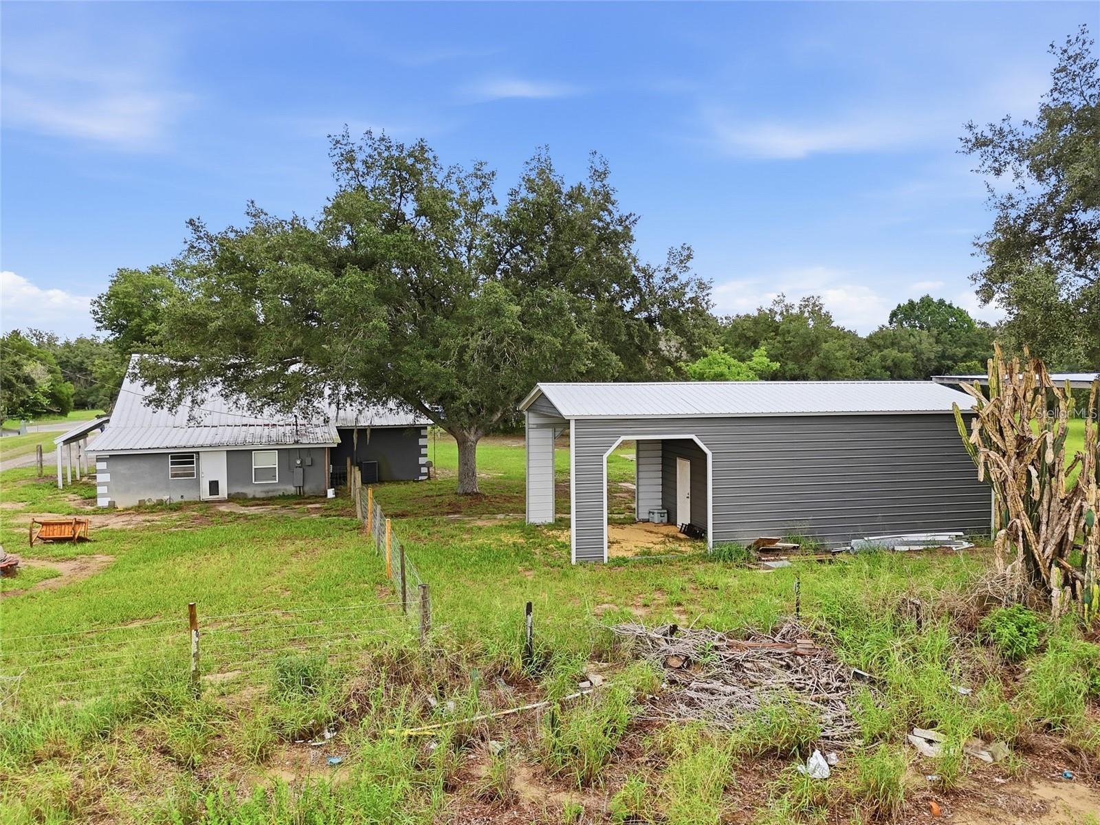 Gray house and large metal outbuilding beneath a mature oak tree on a grassy, fenced property.
