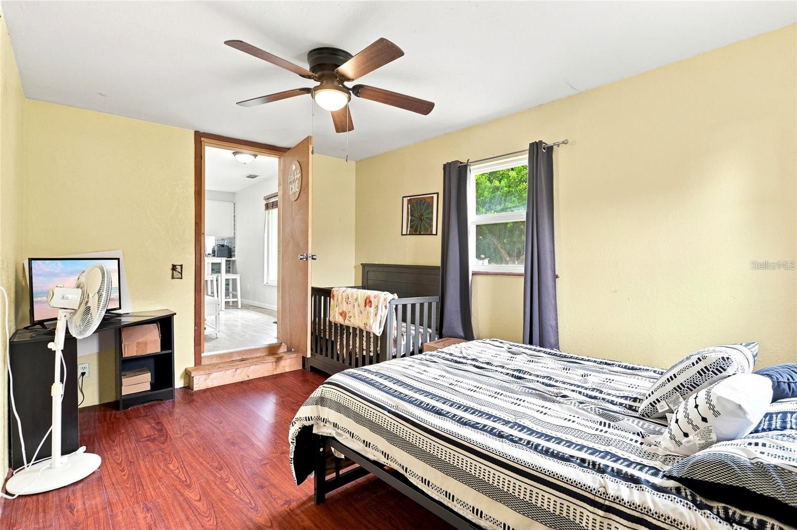 Bedroom with patterned bedding, a crib beside the window, and reddish wood flooring.