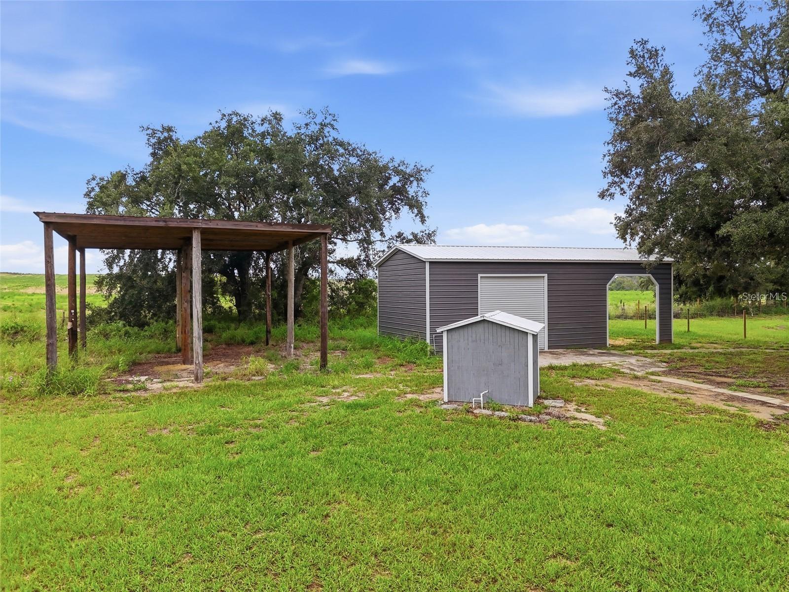 Grassy yard with a gray metal outbuilding, open-sided wooden shelter, and small utility enclosure.