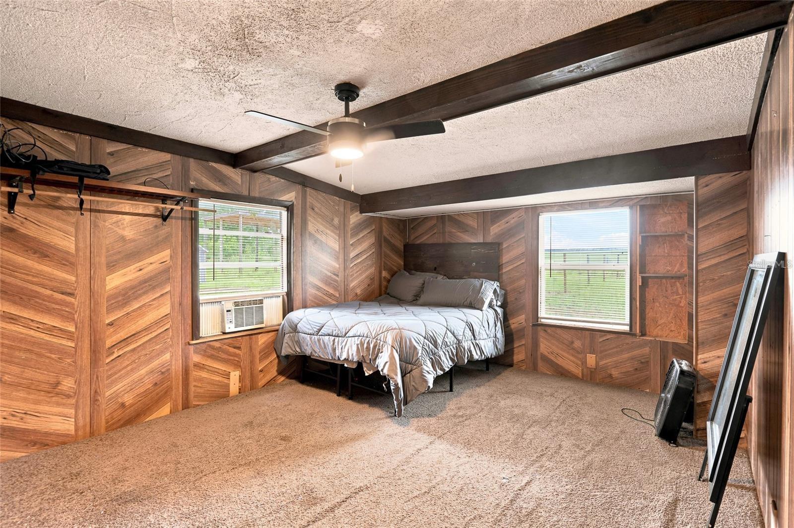 Bedroom with wood-paneled walls, exposed ceiling beams, and a bed between two windows.