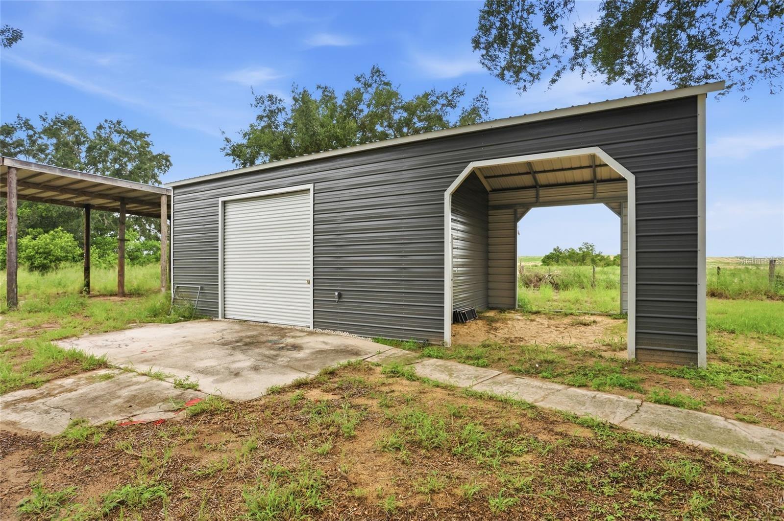 Dark gray metal outbuilding with a white roll-up door, open bay, and adjoining covered carport.