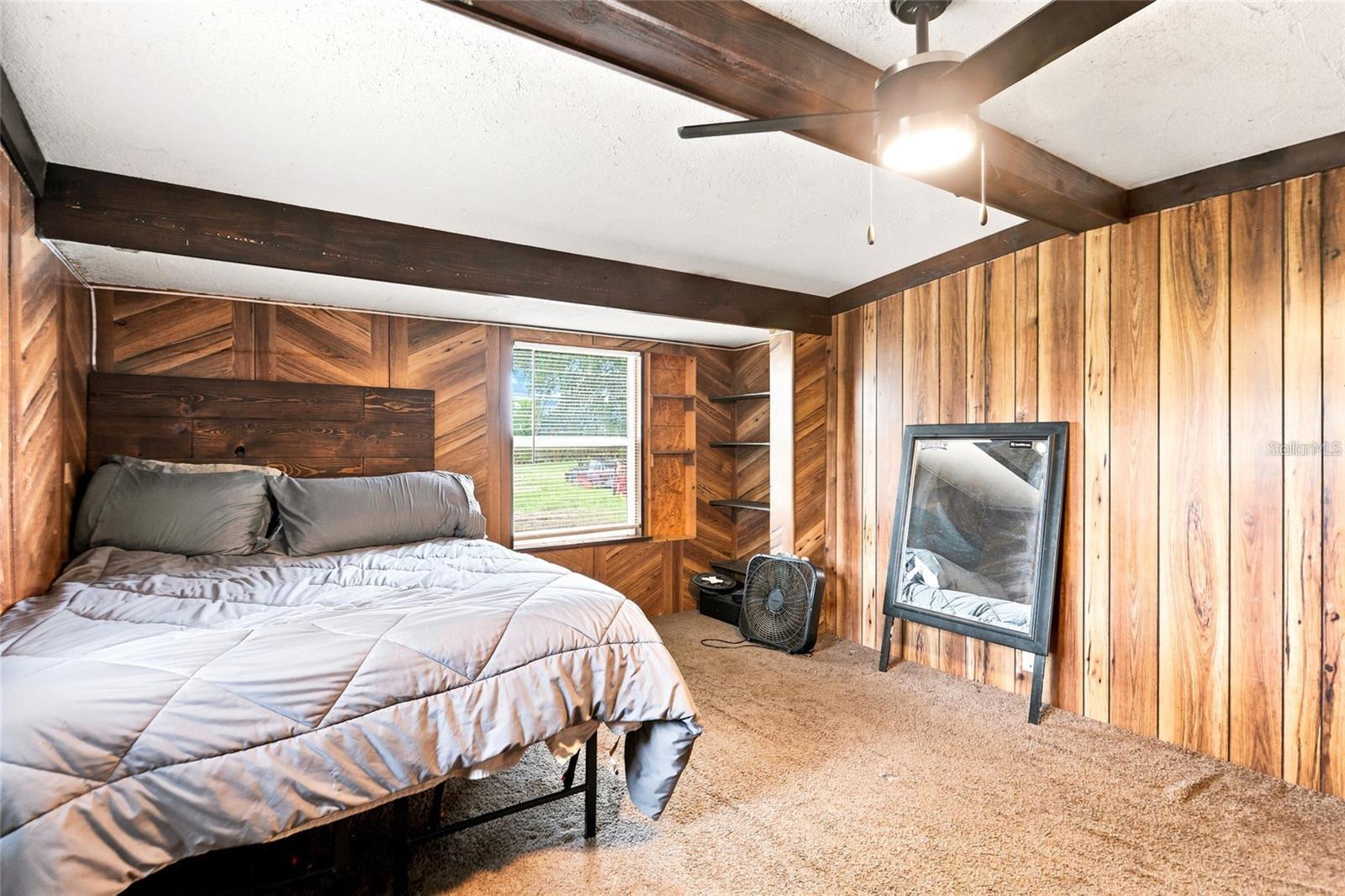 Bedroom with wood-paneled walls, exposed ceiling beams, and built-in corner shelves.
