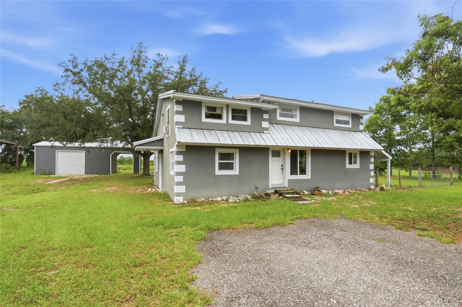 Gray two-story home with a light metal roof, detached outbuilding, and gravel driveway.