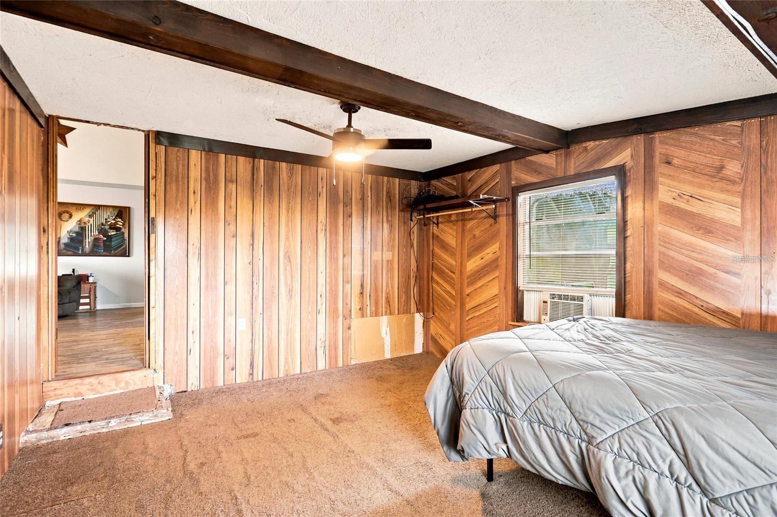 Bedroom with wood-paneled walls, exposed ceiling beams, and a bed beside a window air conditioner.