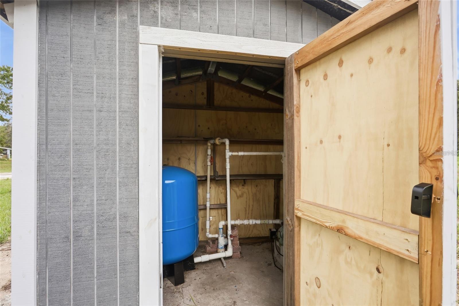 Utility shed interior with a blue pressure tank and exposed plumbing behind an open wooden door.