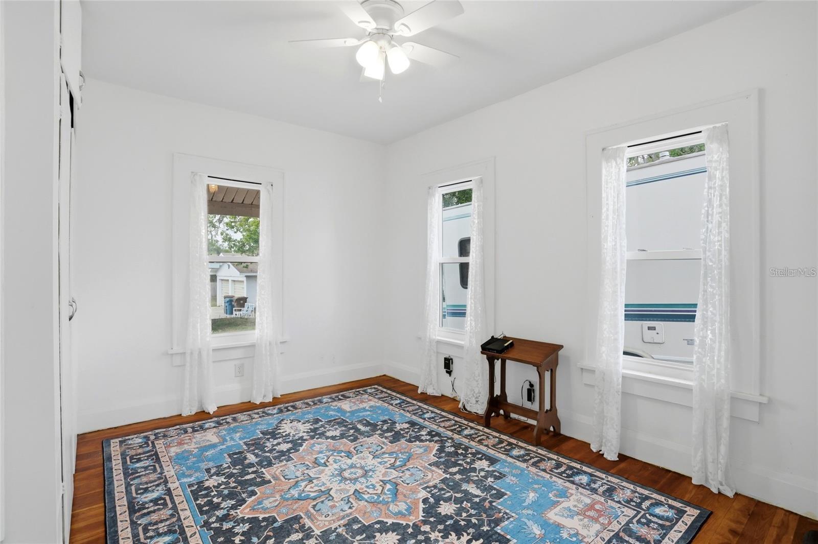 Bright room with hardwood floors, a blue patterned rug, three curtained windows, and a ceiling fan.