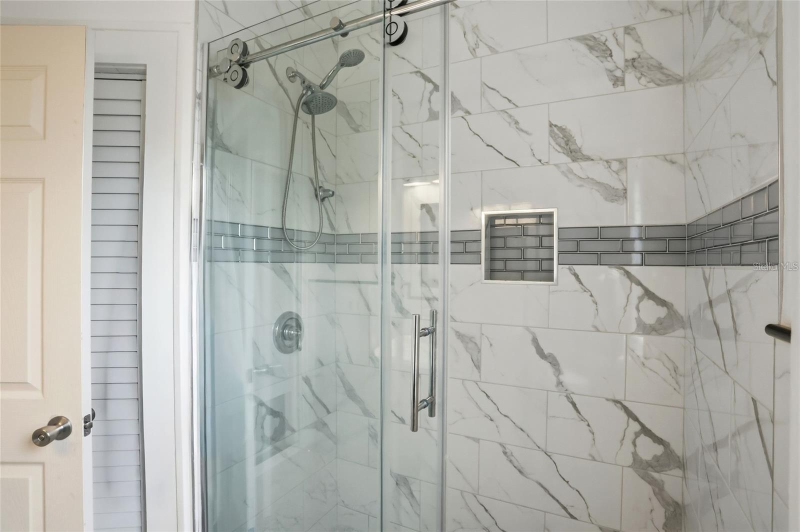 Bathroom shower with glass doors, marble-look tile, gray accent band, and recessed wall niche.