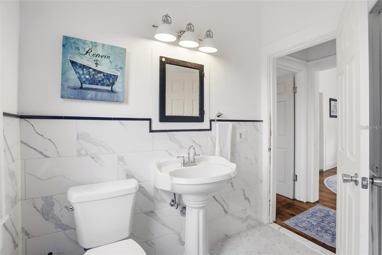 Bathroom with marble-look tile, pedestal sink, and dark-framed mirror.