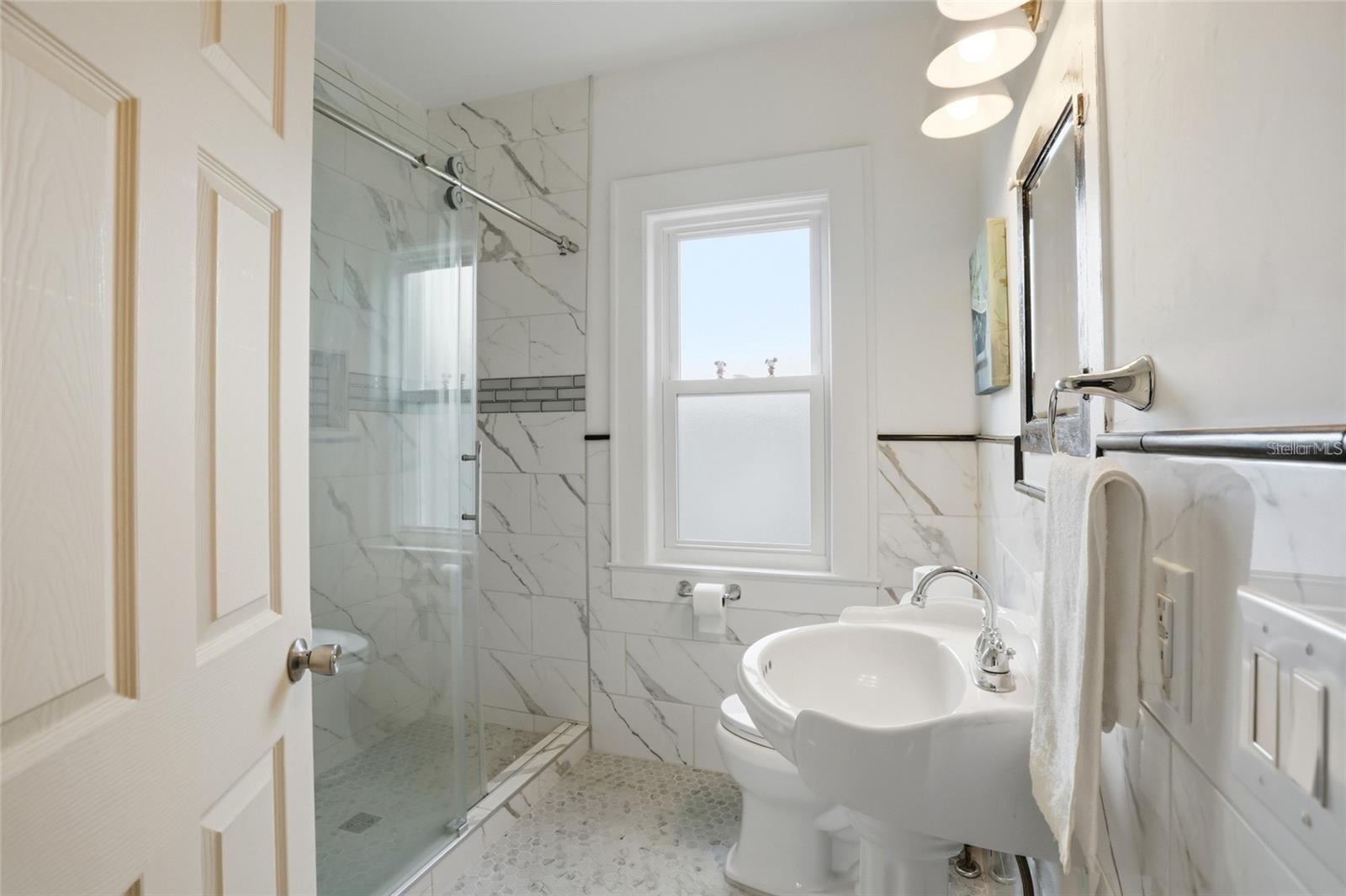 Bright bathroom with a glass shower, marble-look tile, hexagonal floor, and pedestal sink.