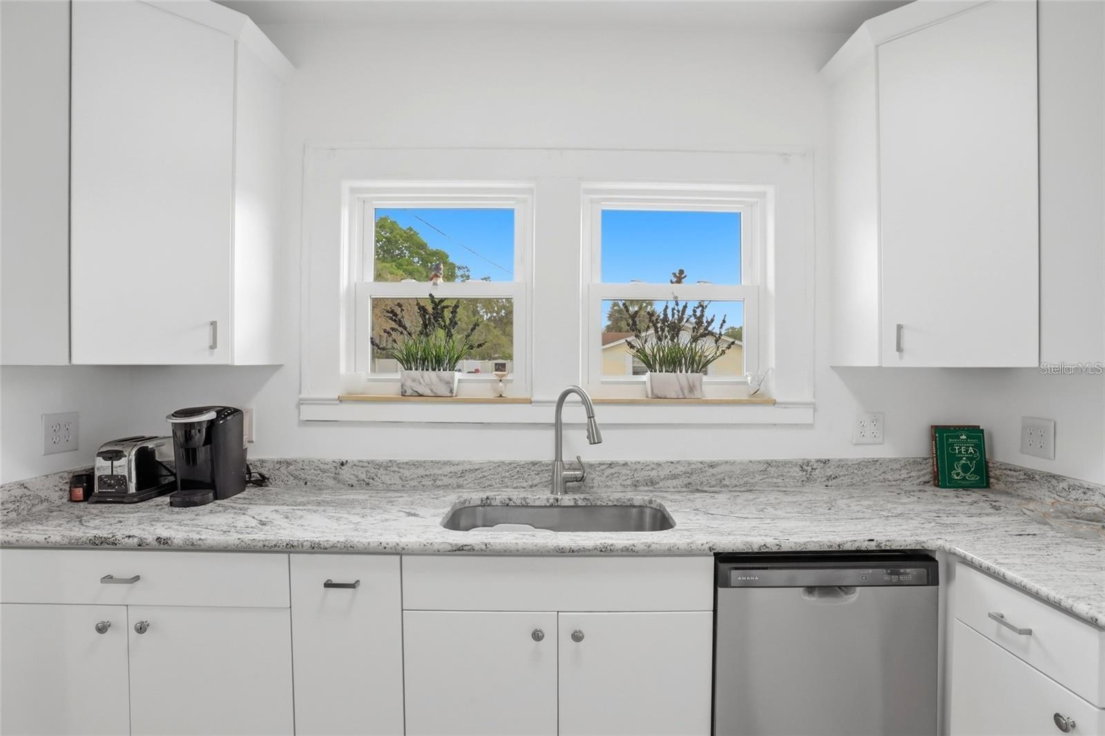 Bright white kitchen with stone countertops, a stainless steel sink, and two windows above the counter.