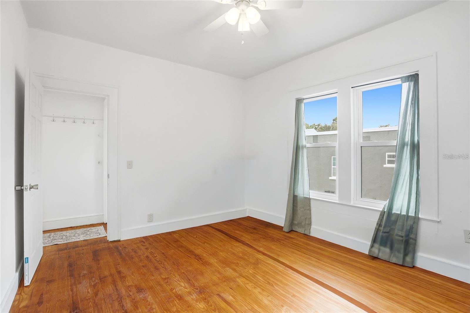 Vacant bedroom with polished hardwood floors, two curtained windows, and a ceiling fan.
