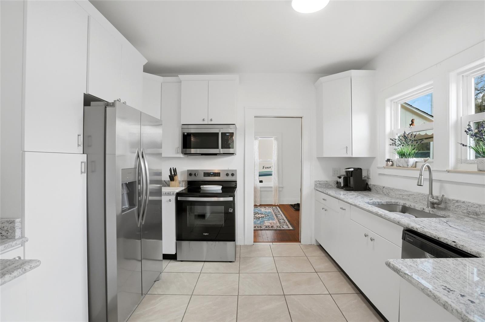 Bright white kitchen with stainless steel appliances, granite counters, and a sink beneath the windows.